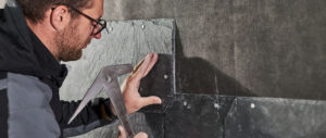 Eine Hand hält eine dunkle Schieferplatte an eine Wand, befestigt mit Nägeln, im Rahmen von Dachdeckerarbeiten.