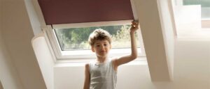 Ein Junge mit lockigen Haaren steht vor einem Fenster und trägt ein graues ärmelloses Shirt.