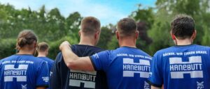 Zwei Männer stehen nebeneinander im Freien, Arm auf der Schulter des anderen, mit Bäumen und blauem Himmel im Hintergrund.