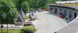 Ein gepflasterter Weg mit angrenzender Straße, im Hintergrund Paletten, Müllcontainer und Bäume, Sommertag.