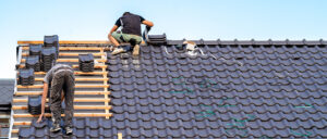 Eine Person arbeitet auf einem Dach mit dunklen Ziegeln, im Hintergrund einige Werkzeuge und Materialien, der Himmel ist klar und blau.