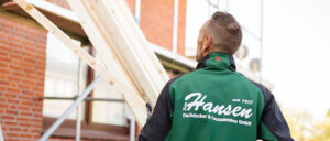Ein Arbeiter trägt grüne Arbeitskleidung und Handschuhe, steht vor einem Holzgerüst mit Ziegelwand im Hintergrund.