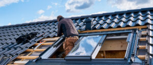Eine Person arbeitet auf einem Dach mit Dachziegeln und einem Dachfenster, Werkzeuge und Anleitungen sind sichtbar, Himmel mit Wolken im Hintergrund.