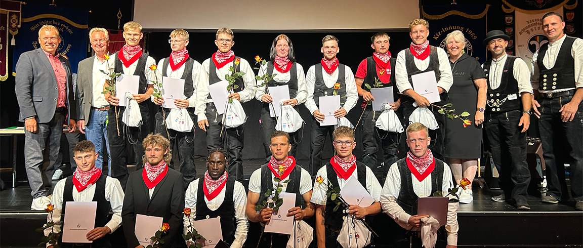 Drei Personen in traditioneller Kleidung halten Rosen und Urkunden, posieren auf einer Bühne und lächeln in die Kamera.