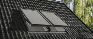 Schrägdach mit dunklen Ziegeln, darauf befinden sich Solarmodule und ein geöffnetes Dachfenster mit Rollläden.
