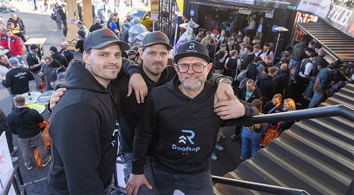 Drei Männer stehen nebeneinander, tragen schwarze Hoodie mit „Rooftop“-Logo, Menschenmenge und Ausstellungsstücke im Hintergrund.