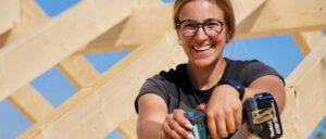 Eine Person mit Brille und Ohrhörern sitzt draußen, Sonnenlicht beleuchtet den Arm, leicht verschwommener Hintergrund aus hellem Holz und blauem Himmel.