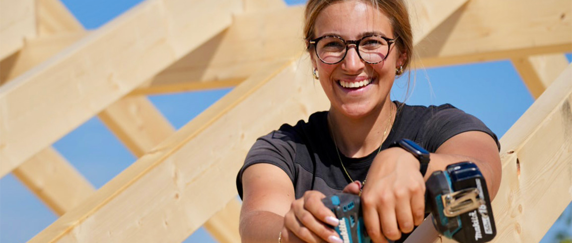 Eine Person mit Brille und Ohrh&ouml;rern sitzt drau&szlig;en, Sonnenlicht beleuchtet den Arm, leicht verschwommener Hintergrund aus hellem Holz und blauem Himmel.