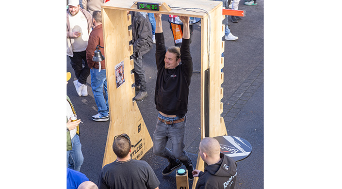 Ein Mann h&auml;ngt an einer Stange, tr&auml;gt Jeans und einen schwarzen Hoodie, Menschen stehen um ihn herum, eine Stoppuhr zeigt eine Zeit an.