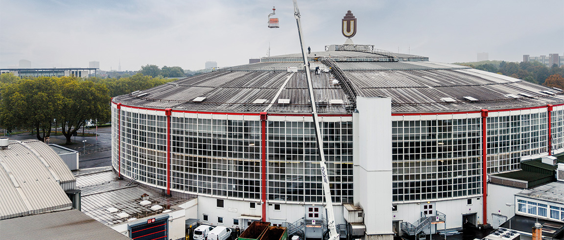 Ein Kran hebt eine Palette mit Baumaterial auf das Dach eines großen Industriegebäudes mit vielen Fenstern.