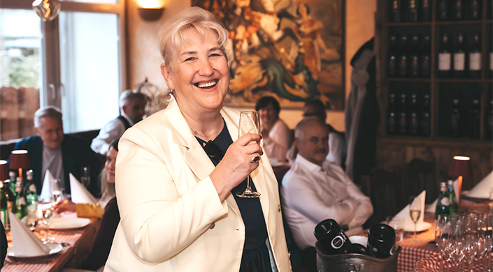 &Auml;ltere Frau mit wei&szlig;em Haar l&auml;chelt und h&auml;lt ein Glas Sekt in der Hand, im Hintergrund sitzen weitere Personen in einem festlichen Raum.