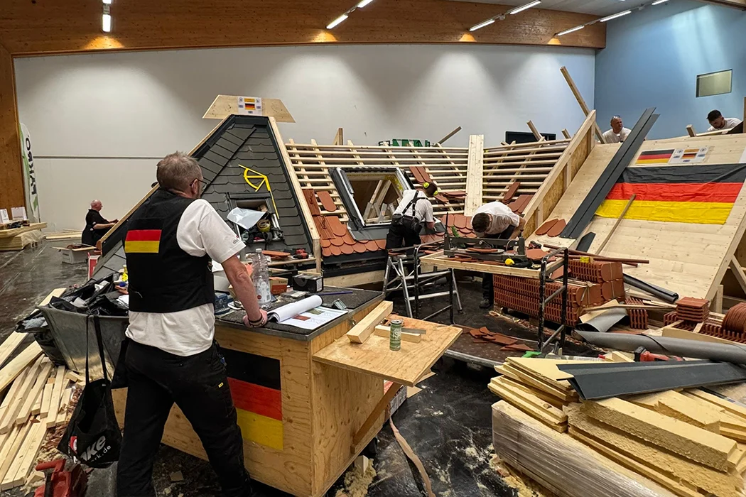 Mehrere Arbeiter bauen in einer Halle an Dachkonstruktionen mit deutschen Flaggen, Holz und Dachziegeln, Arbeitsmaterialien und Werkzeugen sind um sie herum verteilt.