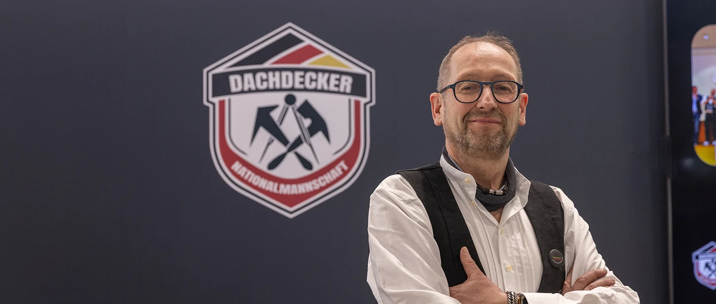 Ausbilder und Trainer Jörg Schmitz, mit Brille, steht vor einem Schild mit der Aufschrift „Dachdecker Nationalmannschaft“ und gekreuzten Werkzeugen darauf.