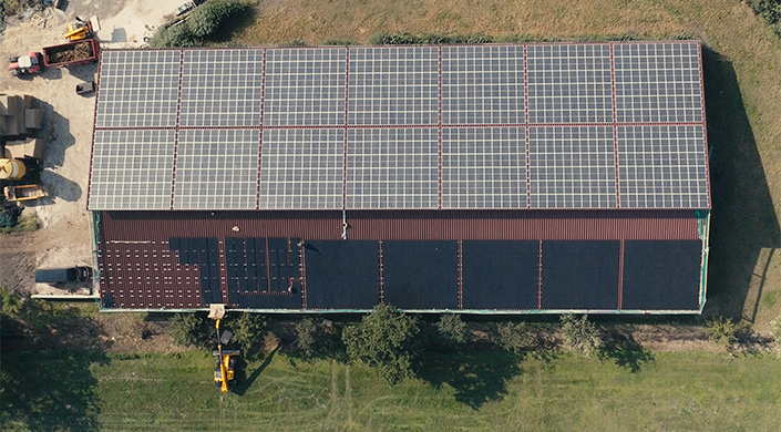 Solarmodule auf dem Dach eines Geb&auml;udes, umgeben von Gras und B&auml;umen, aus der Vogelperspektive fotografiert.