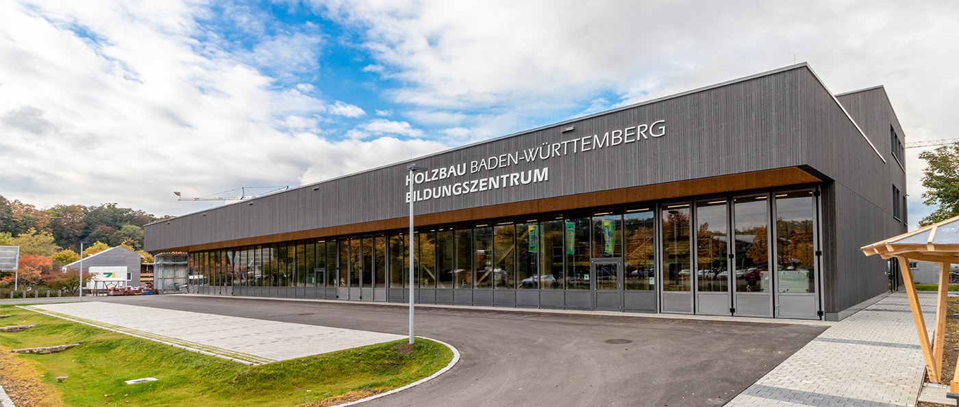 Modernes Gebäude mit großer Glasfassade, darauf steht „Zentrum Holzbau Baden-Württemberg Bildungszentrum“. Im Vordergrund eine Straße, im Hintergrund Bäume und Himmel mit einigen Wolken.