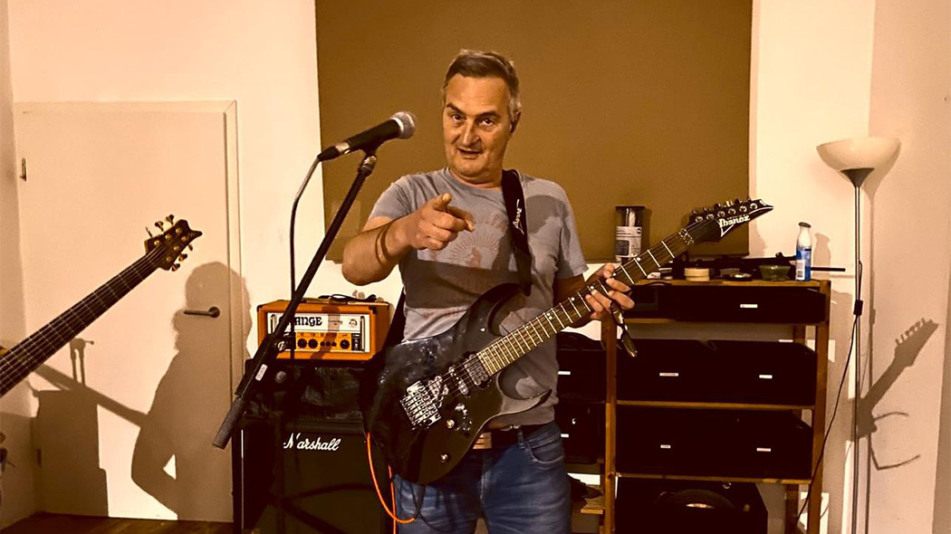 Ein Mann mit Gitarre zeigt in die Kamera und tr&auml;gt ein graues T-Shirt.