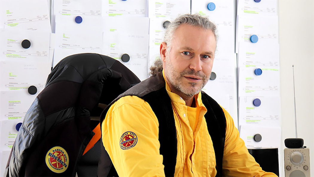Ein Mann mit grauem Haar, tr&auml;gt ein gelbes Hemd mit Logo und eine schwarze Weste, im Hintergrund befinden sich Bl&auml;tter mit Text.
