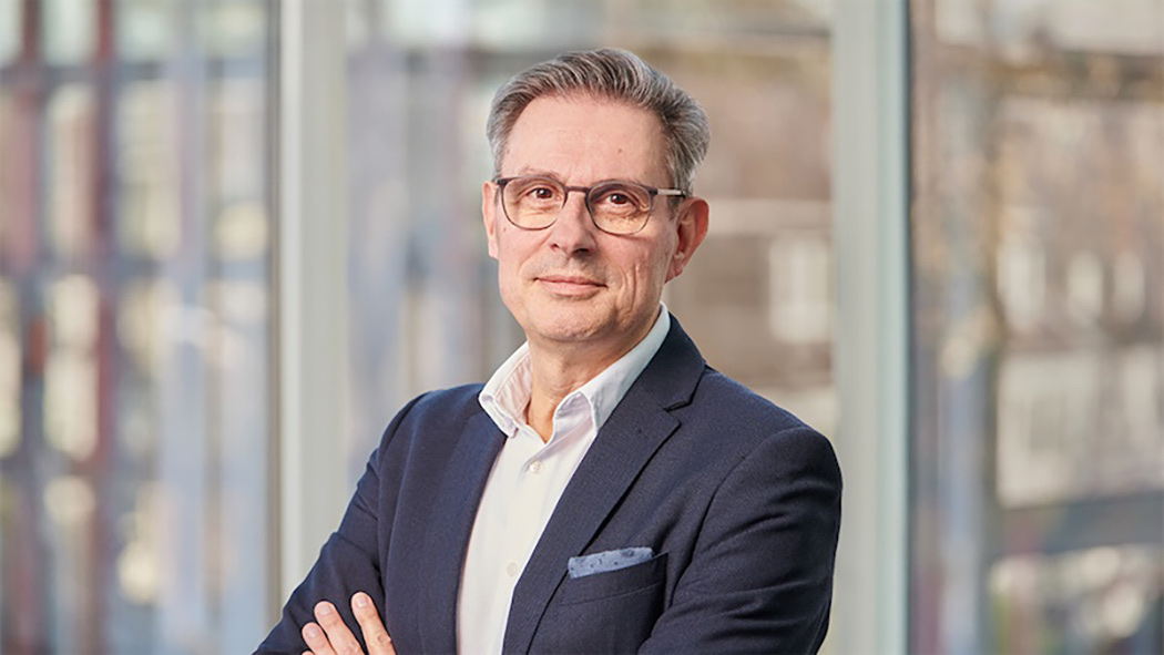 Ein Mann mit Brille, hellem Hemd und blauem Anzug l&auml;chelt leicht in die Kamera.