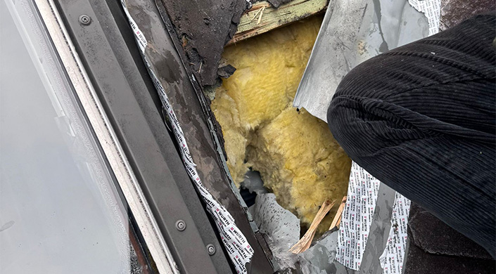 Nahaufnahme von gelber Isolierung, die durch besch&auml;digtes Dachmaterial freigelegt wurde, umgeben von Holz und anderen Baumaterialien.