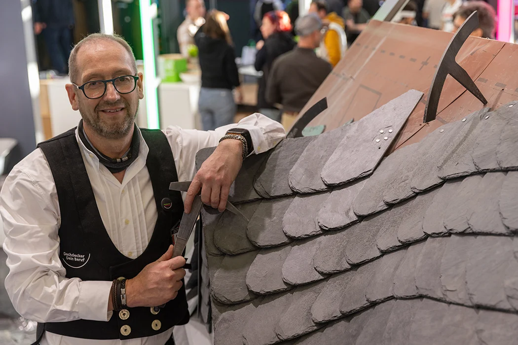 Ein Mann in traditioneller Handwerkerkleidung arbeitet an einem Schieferdachmodell, im Hintergrund sind mehrere Personen in einer Messe- oder Ausstellungshalle.