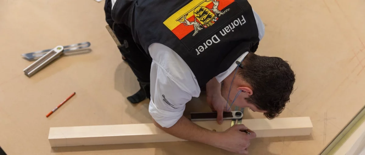 Zimmerer Florian Dorer misst Holz mit einem Messwerkzeug, trägt eine schwarze Weste mit einem Wappen und Namen, arbeitet konzentriert auf dem Boden der Messehalle beim öffentlichen Training auf der Dach+Holz 2026 in Köln.
