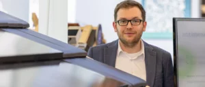 Jan Redecker mit Brille und Bart steht in der Messehalle vor einem PV-Modell neben einem Bildschirm und blickt in die Kamera.