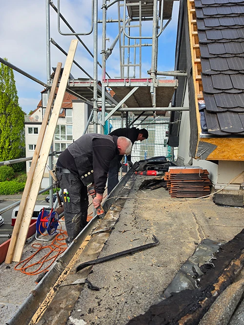 Zwei Handwerker arbeiten auf einem Ger&uuml;st an einem Dach, umgeben von Werkzeugen und Baumaterialien, w&auml;hrend sie Renovierungsarbeiten durchf&uuml;hren.