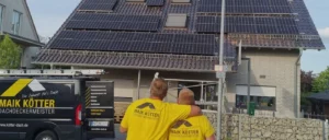 Zwei Dachdecker in gelben Shirts schauen auf ein Haus mit vielen Solarpanels, daneben steht ein Firmenwagen von Maik Kötter Dachdeckerei.