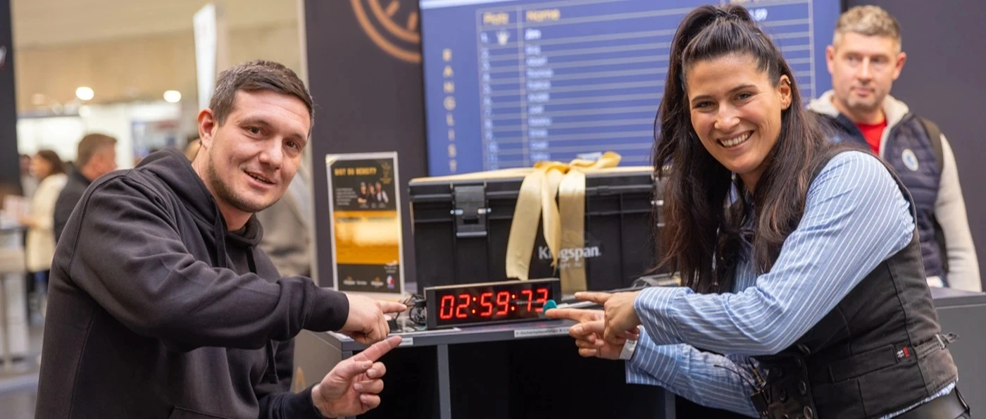 Sonja Theisen zeigt mit einem Teilnehmer der Challenge bei Kingspan auf der Messe Dach+Holz 2026 in Köln auf eine digitale Uhr mit der Zeit 02:59:77, im Hintergrund befinden sich weitere Menschen und eine Rangliste auf einem Bildschirm.