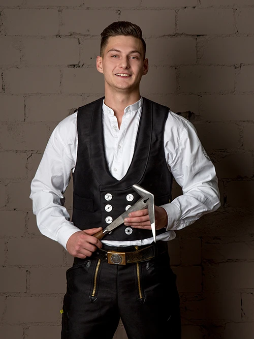 Ein junger Mann in traditioneller Zimmermannskleidung h&auml;lt ein Zimmermannswinkel vor einer gemauerten Wand.
