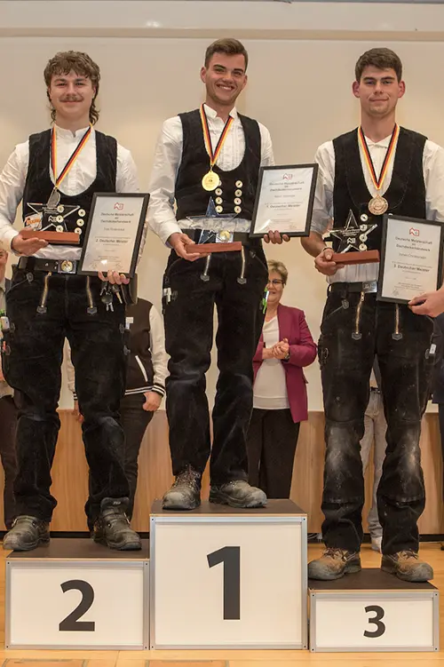 Junger Mann in traditioneller Handwerkerkleidung steht auf einem Siegerpodest, hält eine Medaille, eine Urkunde und eine Trophäe, lächelt stolz.