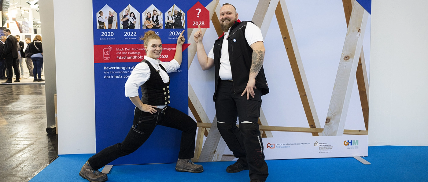 Ein lächelnder Mann zeigt auf ein rotes Schild mit einem Fragezeichen und der Zahl 2028, im Hintergrund Holzbalken.