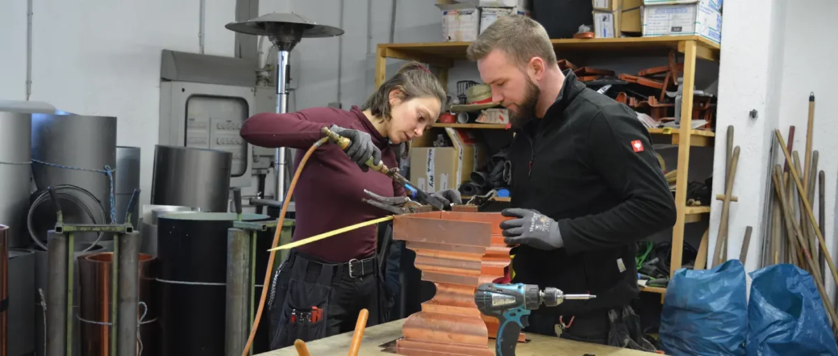 Zwei Personen arbeiten in einer Werkstatt, konzentrieren sich auf ein Metallstück, verwenden Werkzeuge und tragen Arbeitskleidung. Im Hintergrund stehen Regale und Metallrollen.