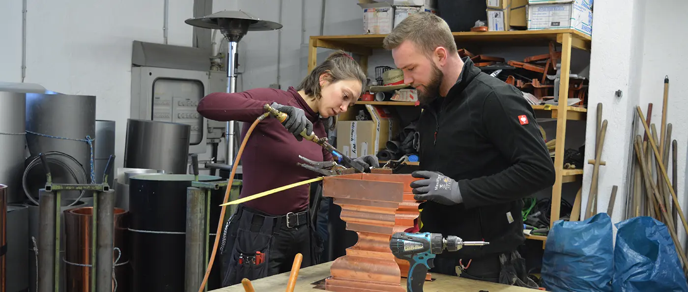 Zwei Personen arbeiten in einer Werkstatt, konzentrieren sich auf ein Metallstück, verwenden Werkzeuge und tragen Arbeitskleidung. Im Hintergrund stehen Regale und Metallrollen.