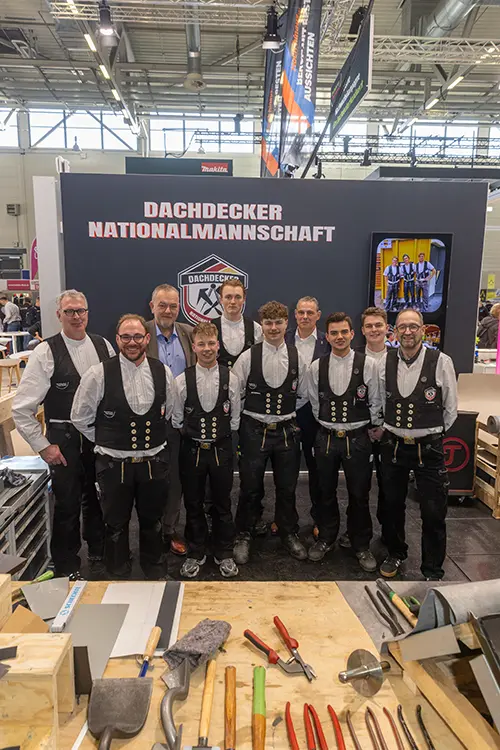 Eine Gruppe von Männern in traditioneller Handwerkerkleidung steht vor einem Stand mit dem Schriftzug „Dachdecker Nationalmannschaft“. Im Vordergrund liegt Werkzeug auf einem Tisch.