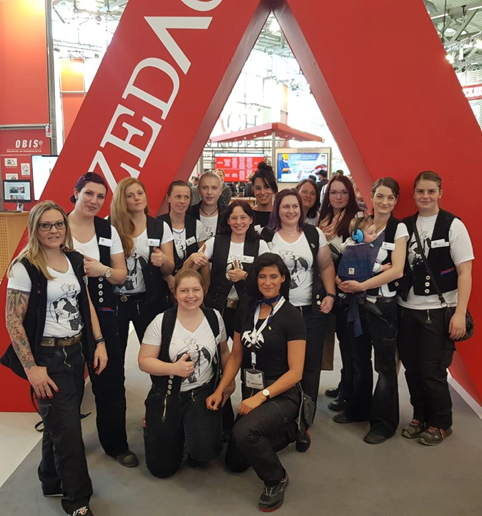 Eine Gruppe von Frauen in einheitlicher Arbeitskleidung posiert vor einem großen roten Zelt auf einer Messe oder Veranstaltung.