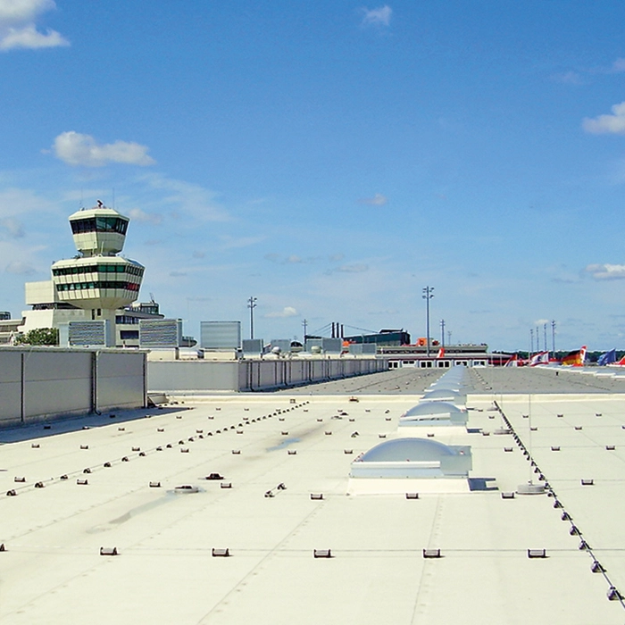 Ein Flughafenterminal mit Kontrollturm im Hintergrund, Flugzeuge sind auf dem Rollfeld sichtbar, der Himmel ist klar und blau mit wenigen Wolken.