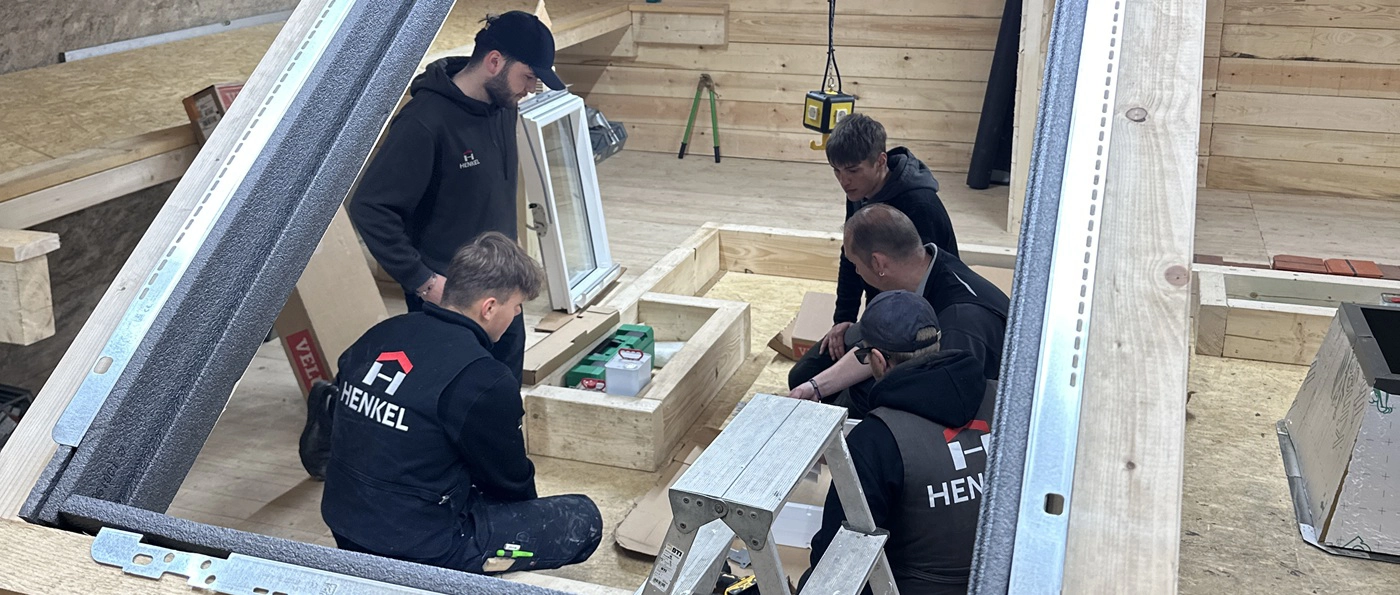 Fünf Handwerker arbeiten gemeinsam an einem Holzrahmen und einem Fenster in einer Werkstattumgebung.