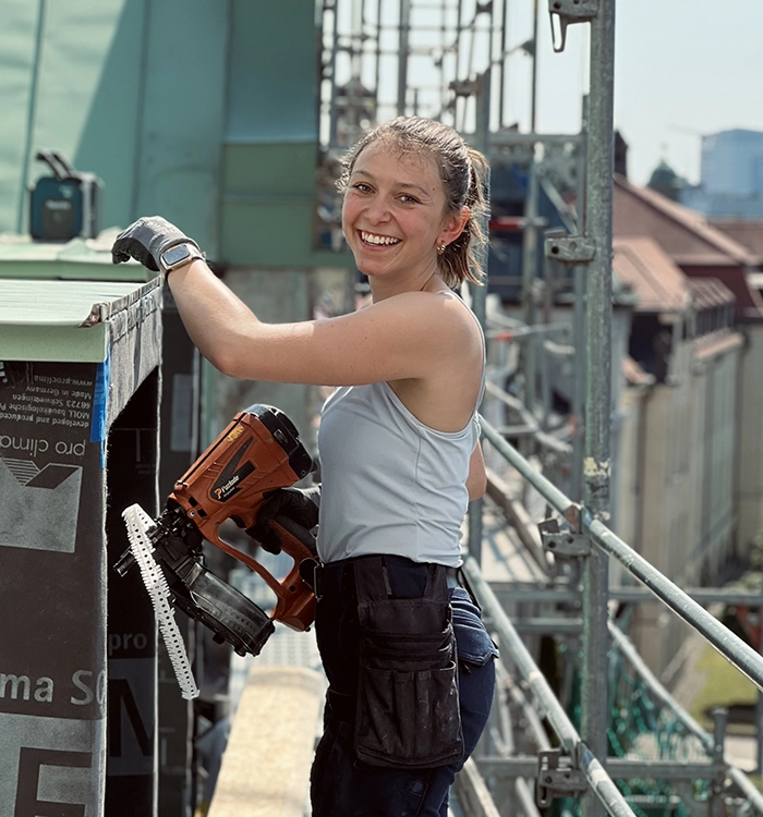 Junge Frau auf einer Baustelle, hält eine Nagelpistole und lächelt, trägt Arbeitshandschuhe und Werkzeugtasche, arbeitet auf einem Gerüst im Freien.