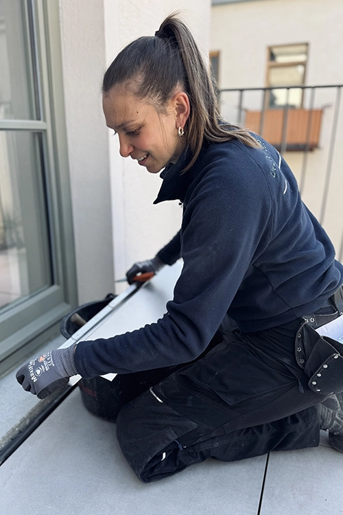 Eine Frau in Arbeitskleidung kniet auf einer Terrasse und misst mit einem Werkzeug eine Fliese ab.