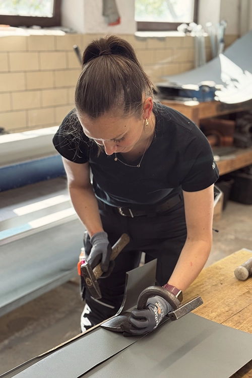 Eine Frau in Arbeitskleidung bearbeitet ein Metallblech mit einem Hammer und einer Zange in einer Werkstatt.