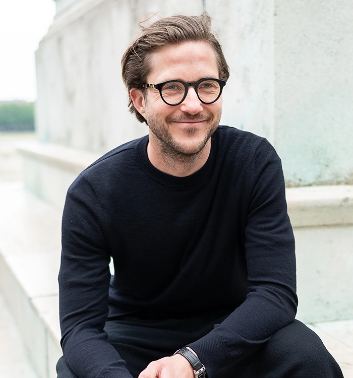 Ein lächelnder Mann mit Brille und dunklem Pullover sitzt draußen auf einer Steintreppe.