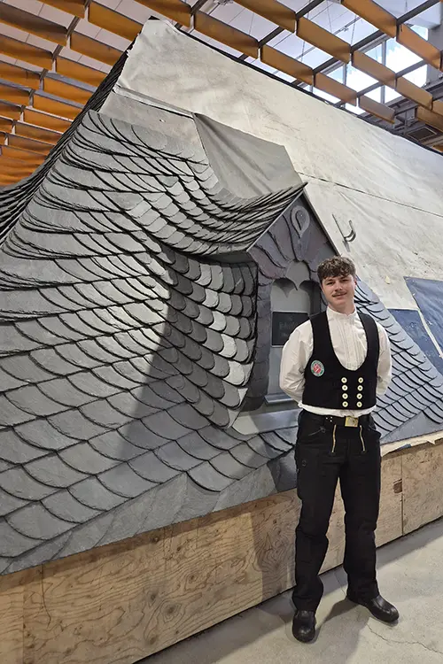 Dach mit kunstvoll angeordneten Schieferziegeln, die ein Muster aus überlappenden Schuppen bilden, unter einer teilweise abgedeckten Holzkonstruktion.
