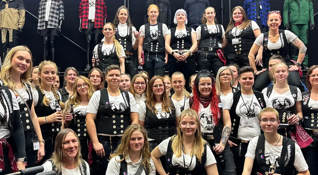 Eine Gruppe von Frauen in traditioneller Handwerkskleidung posiert gemeinsam vor einer Reihe von Schaufensterpuppen mit Arbeitskleidung.