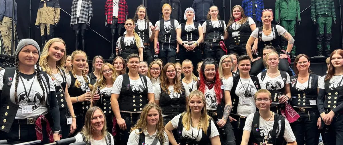 Eine Gruppe von Frauen in traditioneller Handwerkskleidung posiert gemeinsam vor einer Reihe von Schaufensterpuppen mit Arbeitskleidung.