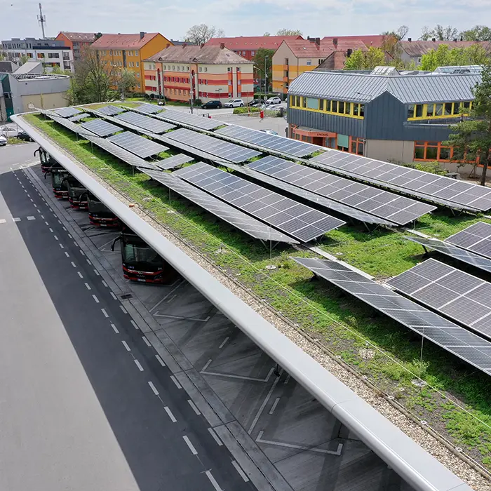 Solardach mit begrüntem Untergrund über einem Busparkplatz, umgeben von Wohngebäuden und einem modernen Gebäude mit gelben Fenstern.