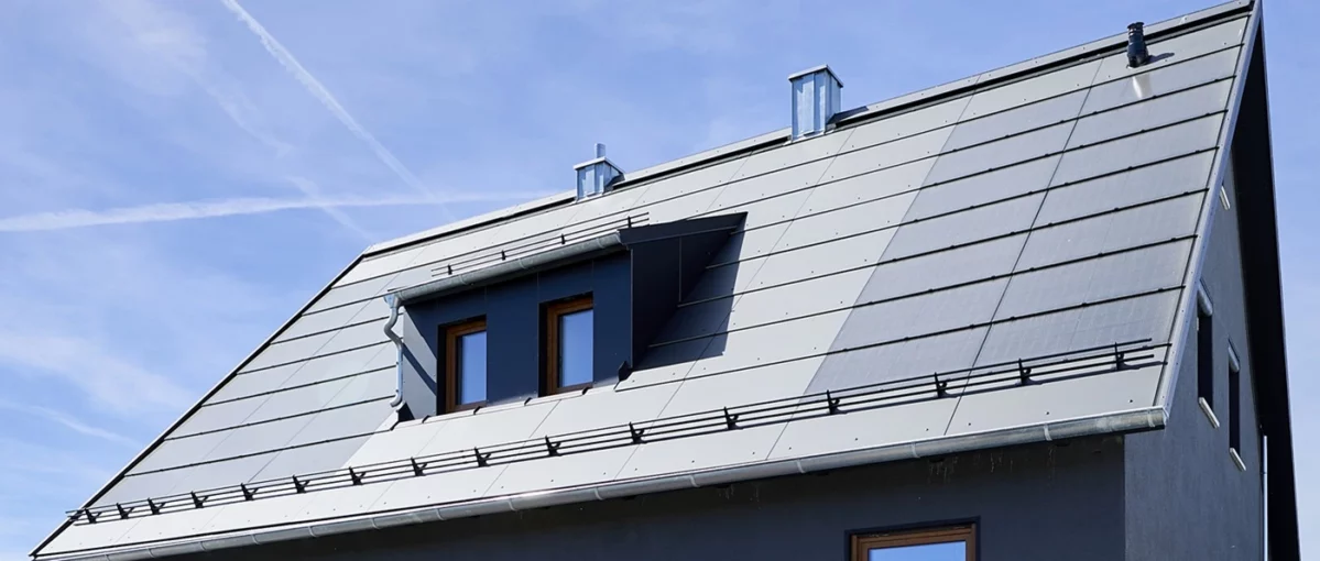 Modernes Schrägdach mit metallischer Abdeckung, Dachfenster und Schneefangsystem unter blauem Himmel.