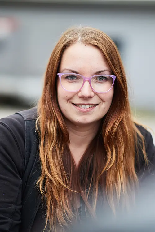 Eine junge Frau mit langen, rotbraunen Haaren und lila Brille lächelt in die Kamera. Sie trägt eine schwarze Jacke und sitzt im Freien.