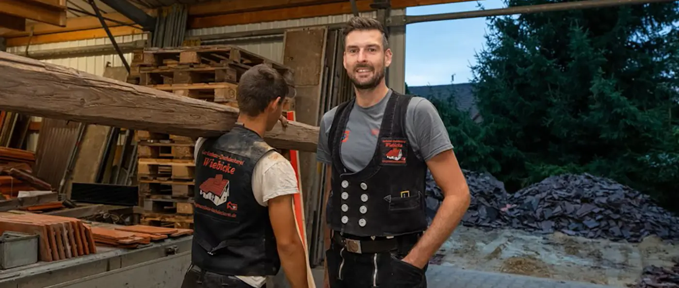 Andreas Wiebicke mit Bart trägt eine traditionelle schwarze Weste mit silbernen Knöpfen und steht vor einem Hintergrund aus Holz und Werkzeugen.