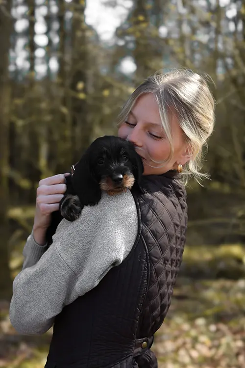 Eine Frau hält einen kleinen schwarzen Hund im Arm und lächelt glücklich, draußen in einer grünen, unscharf dargestellten Umgebung.